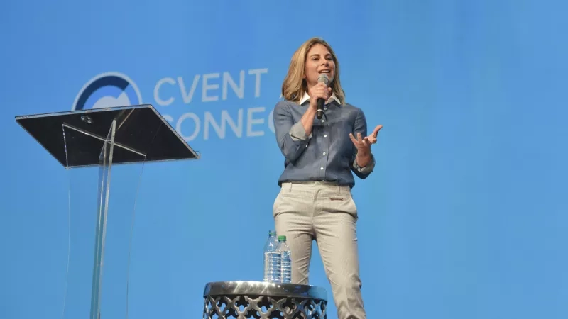 A woman on stage holding a microphone, with a podium and bottles on a table in front of her.