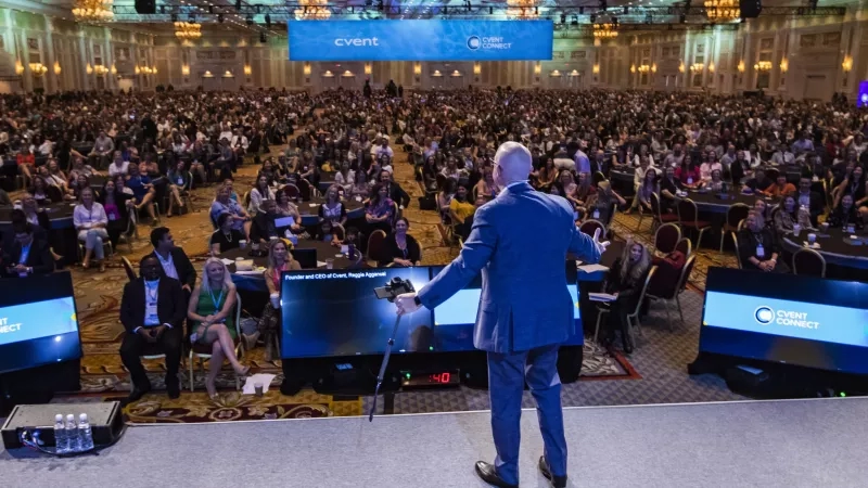 A large crowd in a conference hall with a man standing on stage holding a camera.
