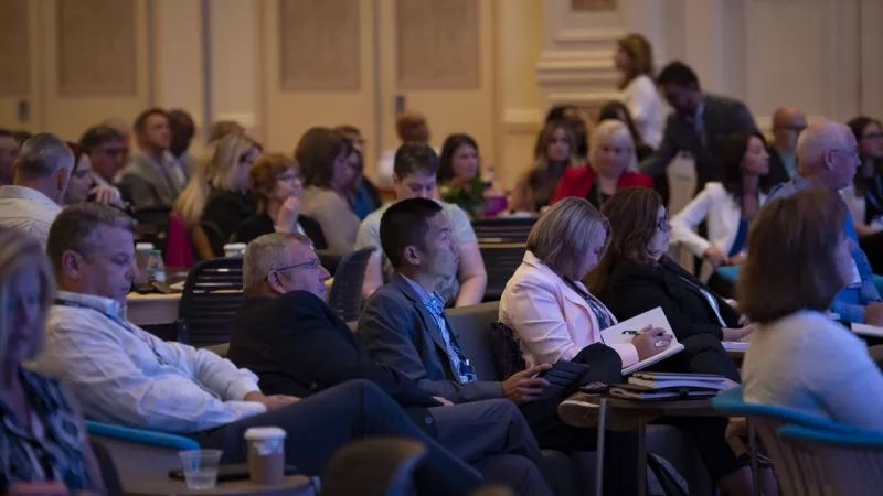 A group of people sit in a conference hall, with some looking ahead and others using their devices.