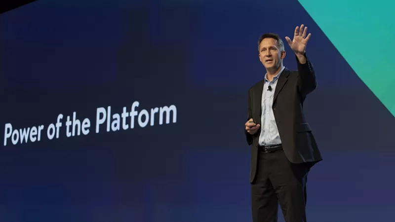 Man standing on stage wearing a suit and holding his hand up in front of a screen.