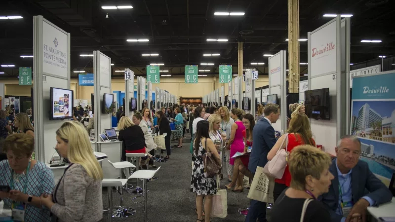 A busy conference hall with many booths, people, and monitors showing text, architecture, and landscaping.