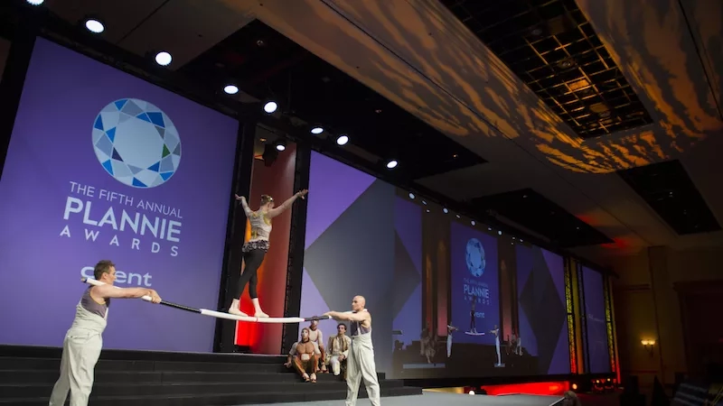 Two people performing a tightrope act on stage at the Fifth Annual Plannie Awards in front of a large screen.