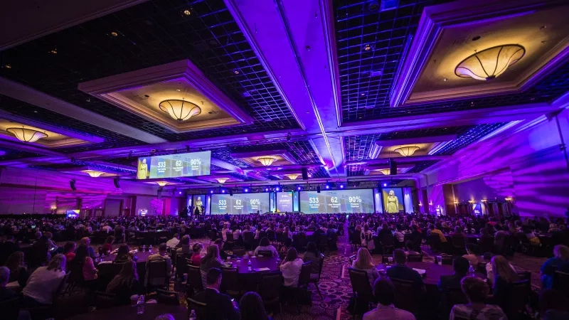 A large conference room with many people seated at tables, purple lighting, and three large screens displaying information.