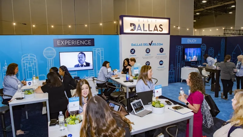 A group of people are sitting at a table in an exhibition hall with a Visit Dallas banner.