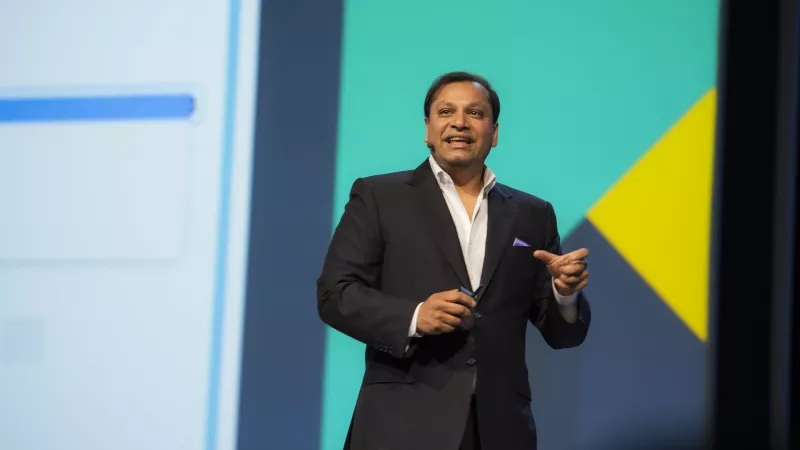 A man wearing a suit standing on stage speaking and gesturing with his hands.