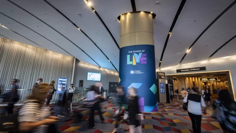 A busy convention center lobby with the words "The Power of Live" on a large blue column.