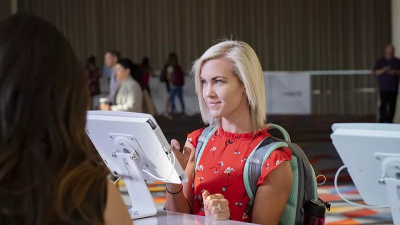 A blonde woman with a floral red dress and a backpack talks to another woman at a convention.
