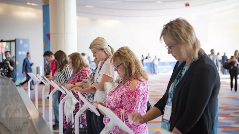 Several people standing in line with electronic tablets, probably in a convention center.