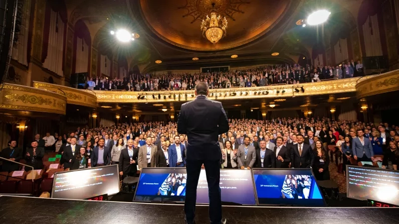 A man stands on stage at a large event with a crowd of people in an auditorium.