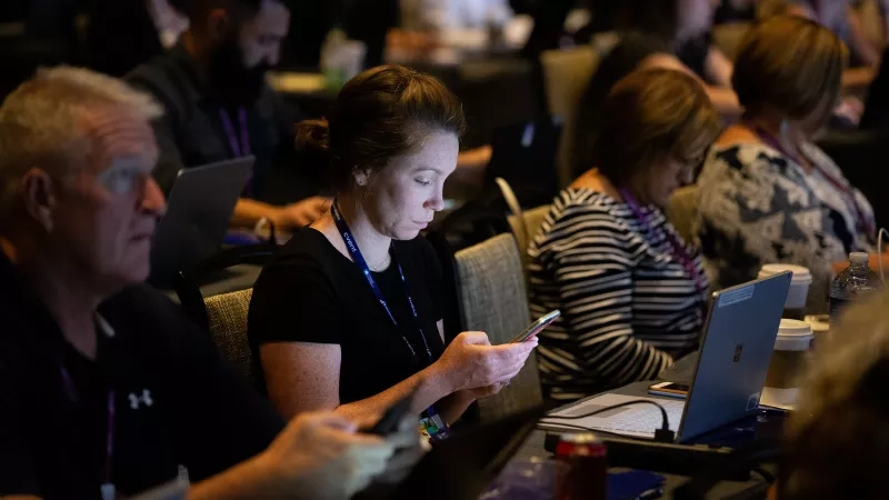 People at a conference, some using their phones and others using laptops, with coffee and a can nearby.