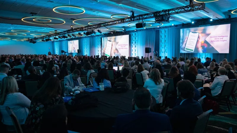 Crowded room with large screens, people seated at tables, and a speaker on stage at an event.
