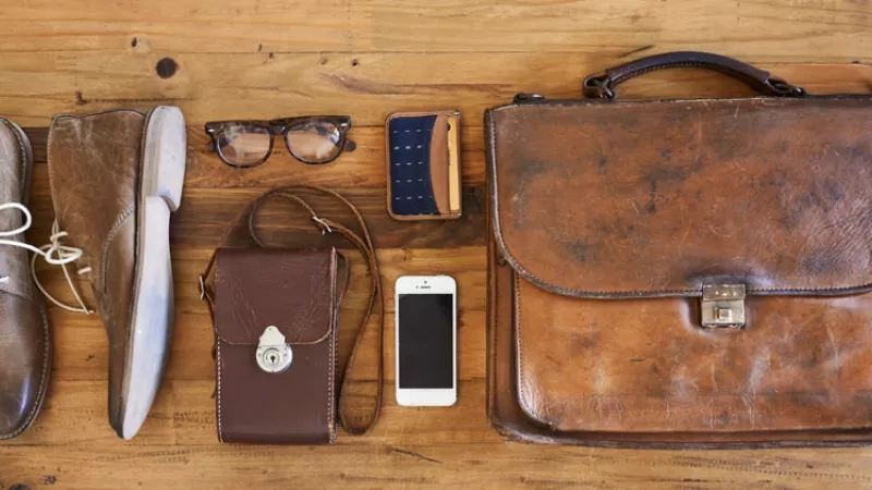 A pair of shoes, eyeglasses, and bags are placed on a wooden surface next to a smartphone.