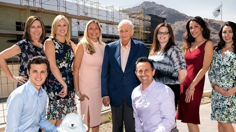 Group of people standing in front of a building, possibly a construction site, with mountains in the background.