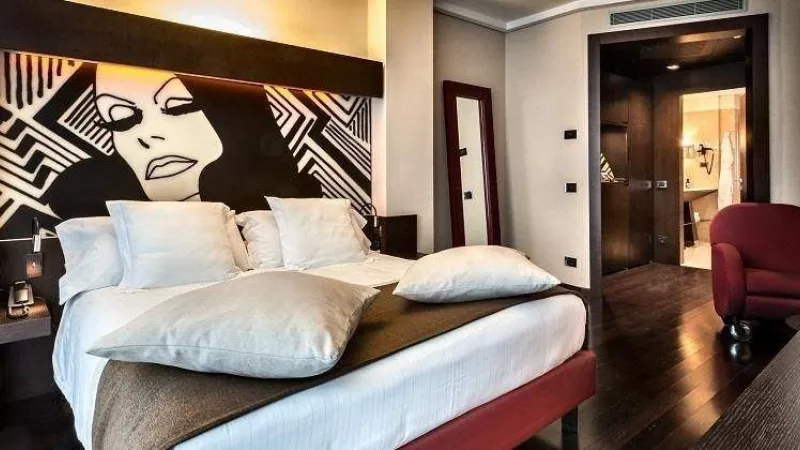 Hotel bedroom with a bed, pillows, and a mural of a woman's face on the wall.