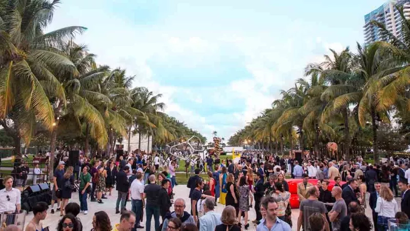 Large crowd of people gathered outside with palm trees, buildings, and a blue sky with clouds.