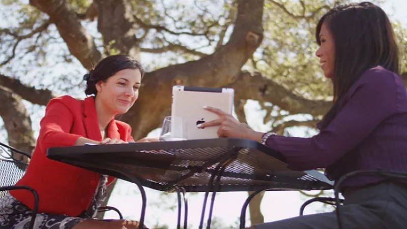 Two women sitting on chairs around a table, holding a tablet in their hands, and smiling.