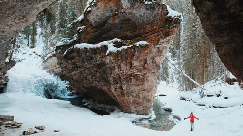 A person stands in the snow at the edge of a large rock formation in Banff National Park.