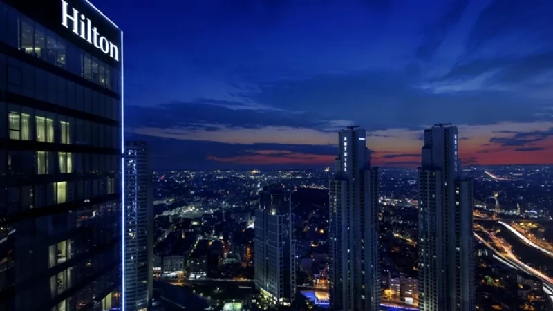 A night cityscape view of the Hilton Istanbul Bomonti hotel with illuminated city lights and buildings.