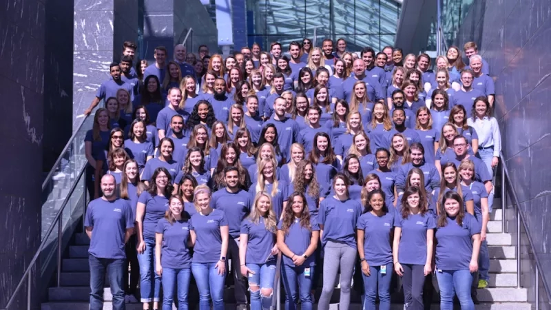 Group of people standing together in front of stairs, wearing the same blue shirt, and smiling for a picture.