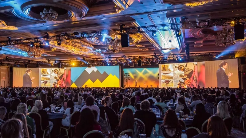 A large crowd of people sit at tables in a large conference room, watching a presentation on a large screen.