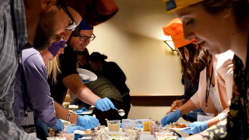 Group of people wearing blue gloves and orange hats in a kitchen, preparing food together.