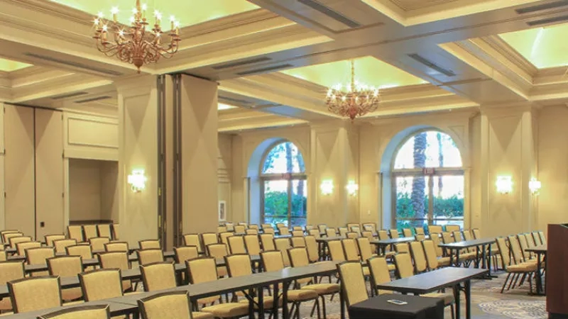 A large conference room with a podium, chairs, desks, chandeliers, and large windows at JW Marriott Desert Springs Resort.