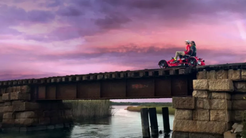 A couple riding a trike across a bridge over a river with a purple sky in the background.