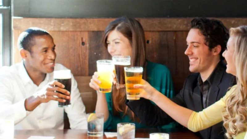 Group of friends drinking beer at a table in a bar with a wooden wall and window in the background.