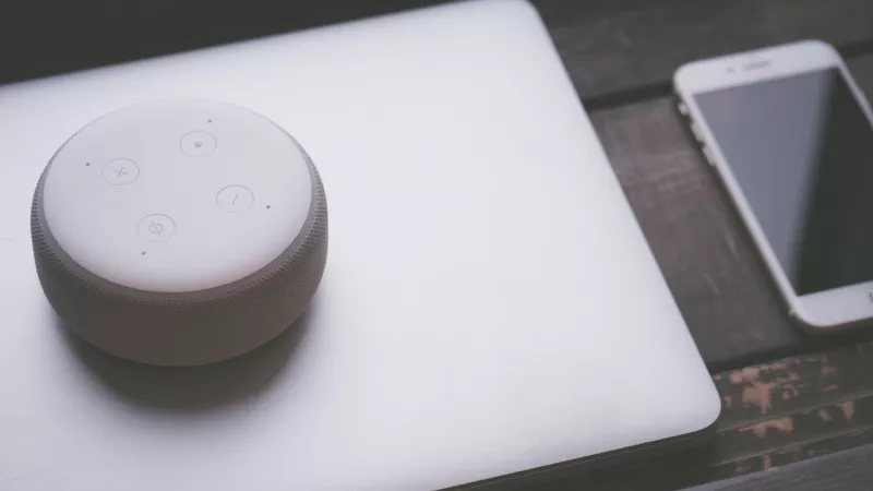 An Amazon Echo Dot sits on top of a laptop computer next to a cell phone.