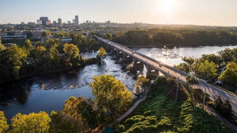Aerial view of Columbia, SC with bridge, river, buildings, and lush greenery under a bright sky.