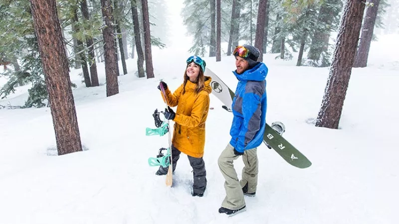 Two people in snow gear walk through a forest holding snowboards in Reno, Nevada.