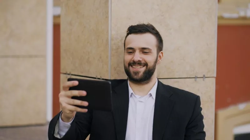 A man with a beard holding a tablet and smiling in front of a building.