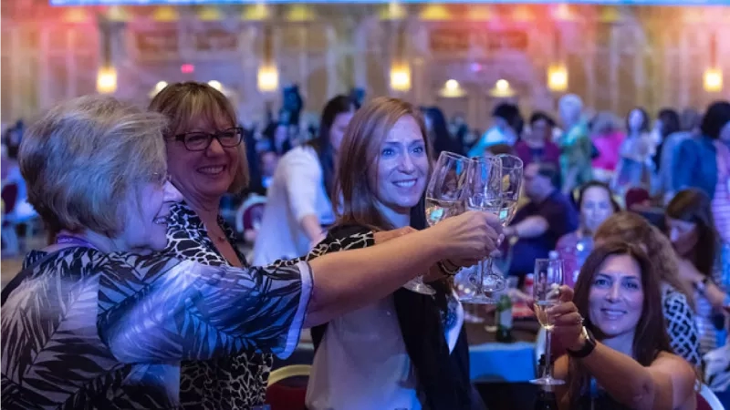 Four women in a banquet hall holding wine glasses with others in the background at the Excellence Awards.