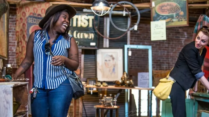 Two women shopping in an antique shop with various items displayed on shelves and tables.