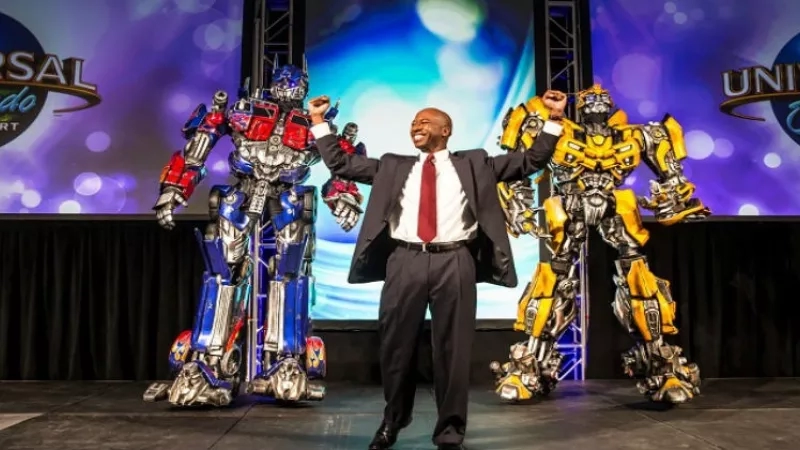 A man stands on stage with two Transformers robots in front of him at Universal Orlando Resort.