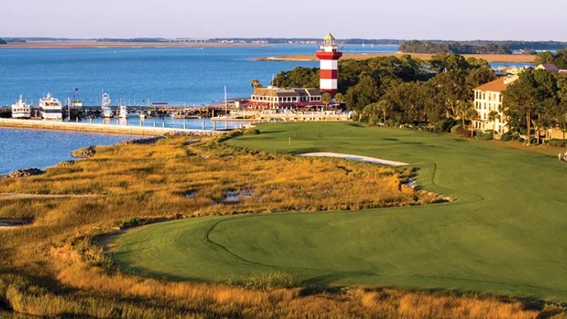 Golf course with a lighthouse in the background and a marina with boats on the left side.