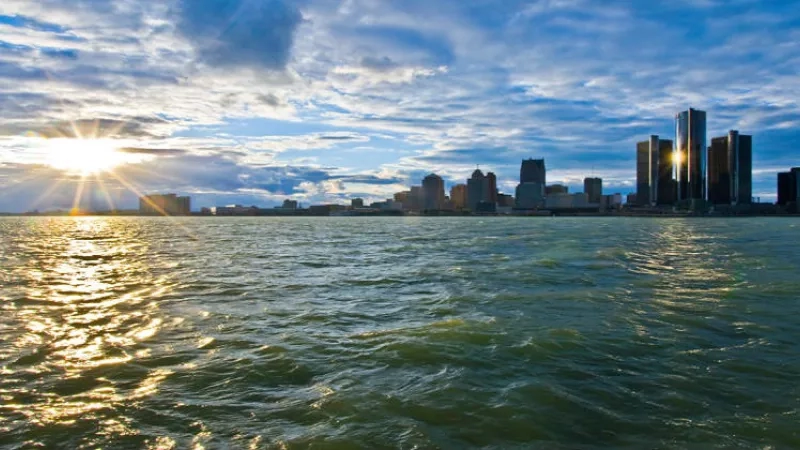 The sun rises behind the Detroit skyline, shining over the water of the Detroit River.