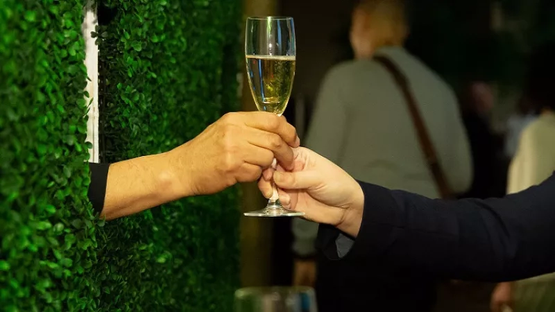 Two people are holding a champagne glass and cheering while other people are standing in the background.