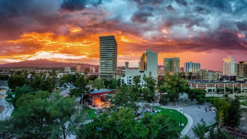Sunset over a city with buildings, trees, a park, and a cloudy sky in the distance.