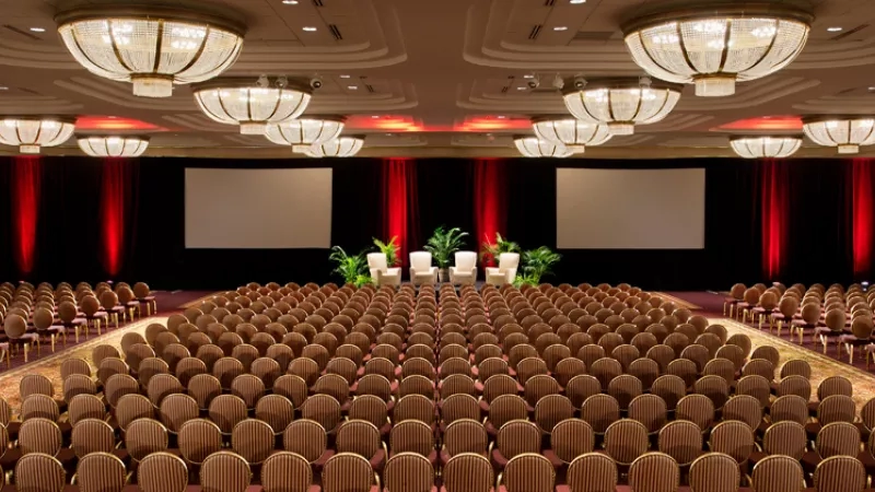 The conference room has chairs, a stage with red curtains, white chairs, plants, and a white screen.