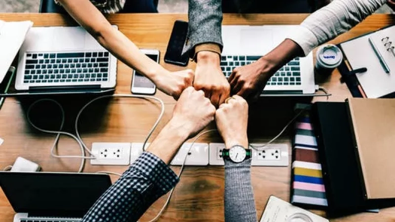 Fist bump meeting of four people sitting around a table with laptops and other items on it.
