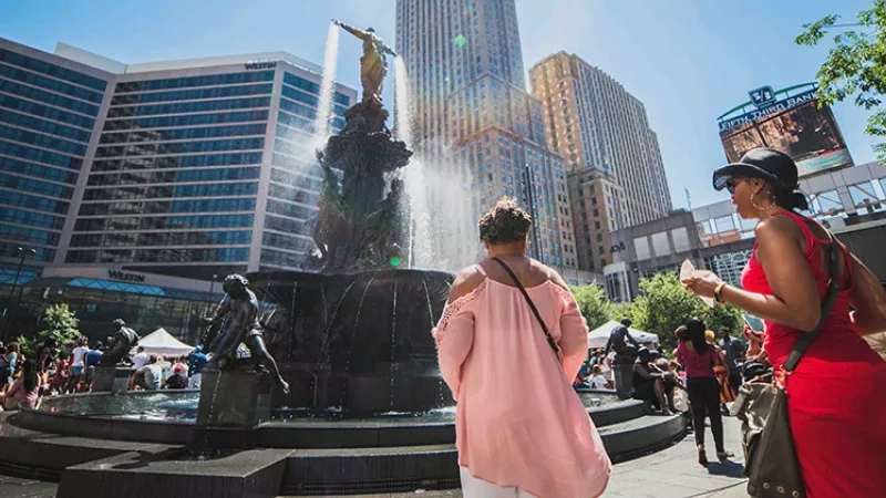 Two women are looking at a fountain in Fountain Square with many people and buildings around.