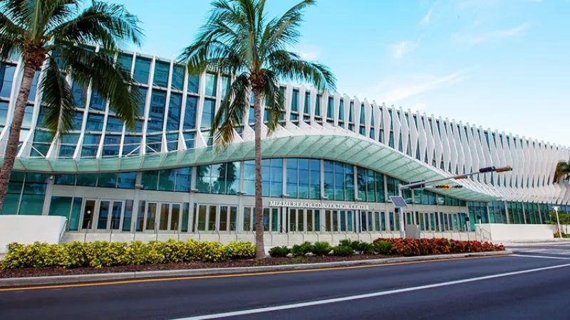 Miami Beach Convention Center with palm trees, glass architecture, and landscaping, under a clear sky.