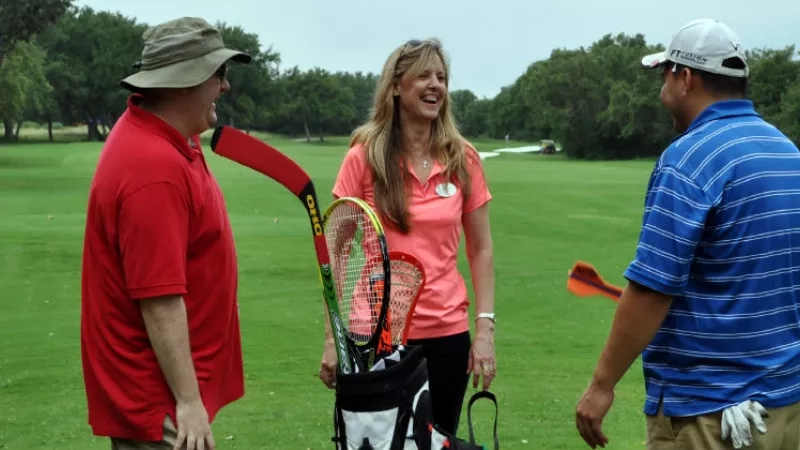 Three people stand on a golf course, two holding hockey sticks, one laughing, with trees in the background.