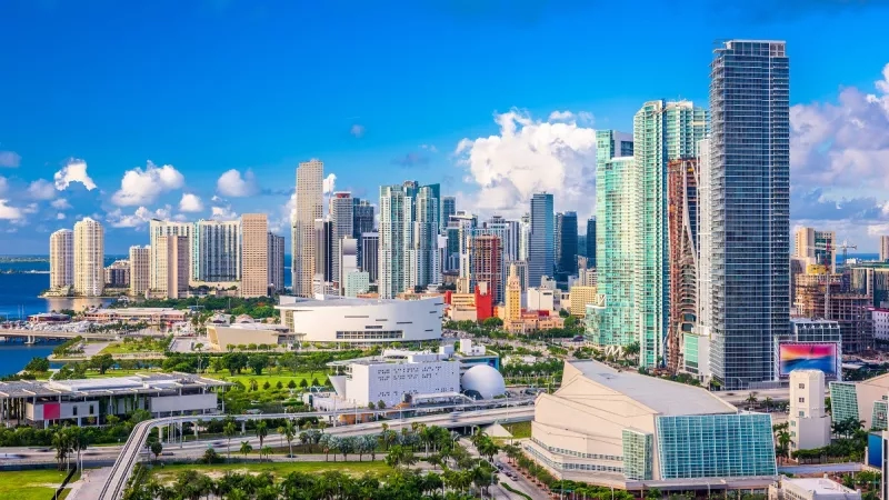 A city skyline with modern architecture, green landscaping, and a vibrant atmosphere under a clear blue sky.