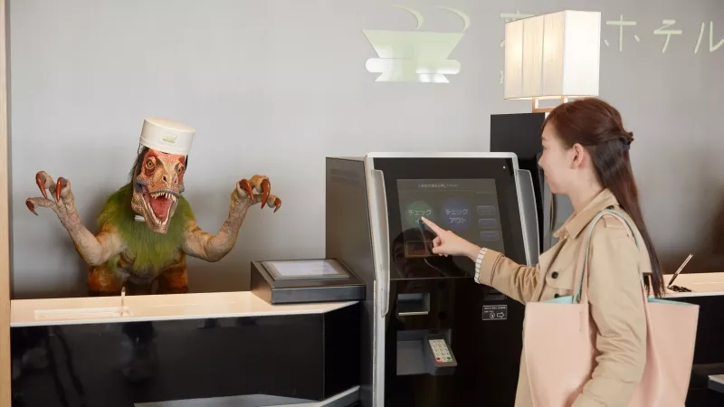 Woman pointing at a machine with a dinosaur mascot behind the counter at Henn-na Hotel.