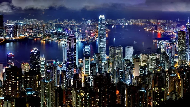 Nighttime aerial view of Hong Kong, showing a bustling cityscape with illuminated skyscrapers and a harbor.