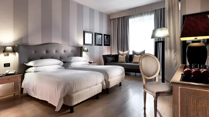 Hotel Ambasciatori's interior view of the bedroom with a bed, lamp, chair, desk, and window.