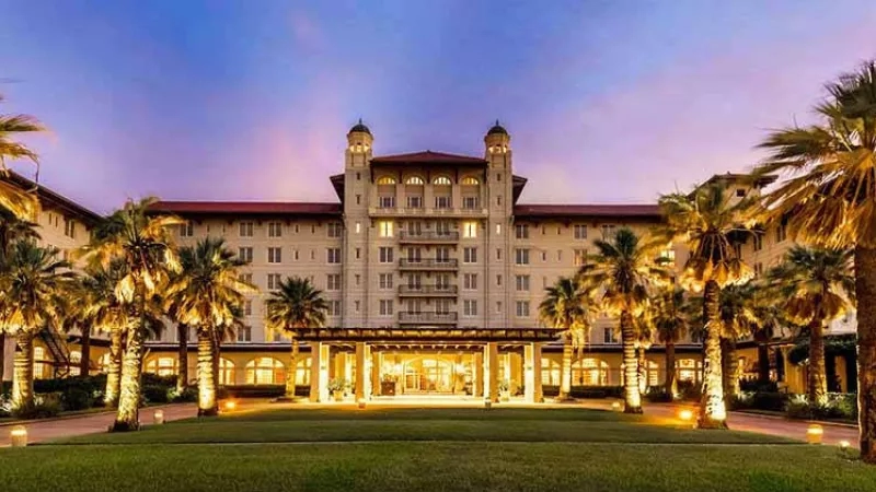 Exterior view of Hotel Galvez with palm trees and lit-up entrance at dusk, highlighting its architectural grandeur.