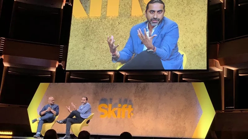 A large screen displaying a man speaking at a conference with an audience in front of him.